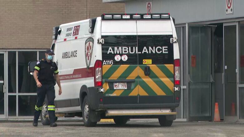 A paramedic wearing a baseball hat and medical mask walks beside an ambulance parked outside a hospital.