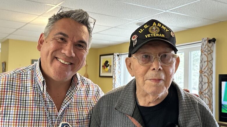 Two man stand together smiling. The man on the left has gray hair and is wearing a striped shirt. The man on the right is wearing a grey sweater and black hat.