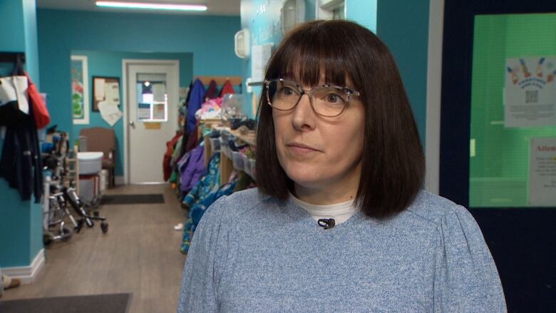 A woman with short brown hair and glasses stands in a day care. There are bright blue walls and children's jackets hanging up behind her. 