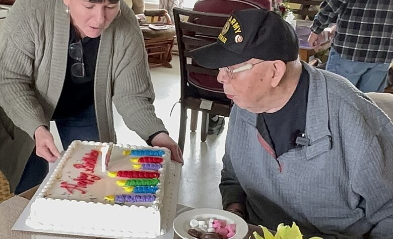 An old man wearing a grey sweater and black baseball cap blows out candles on a large, white birthday cake.