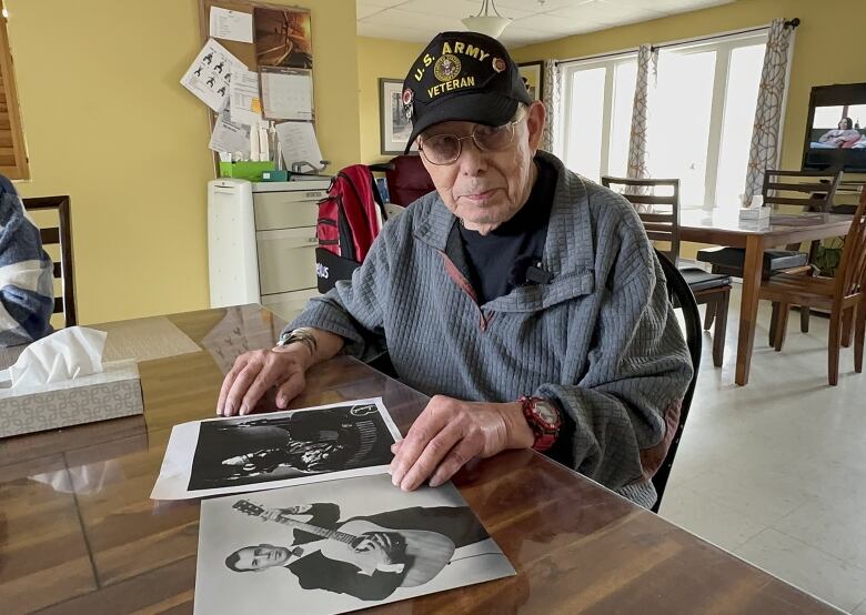 An old man wearing glasses, a grey sweater and a black baseball cap sits at a table with two large, black-and-white photographs on it.