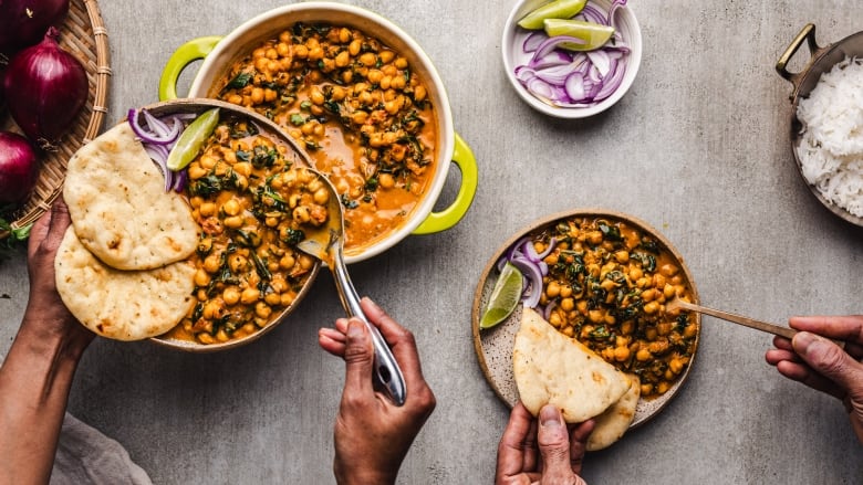 Overhead shot of 2 plates of Chickpea Curry with Spinach. One person's hands are dishing some of the curry onto a plate from a pot. A second person's hands are holding naan and a spoonful of curry over the second plate. 