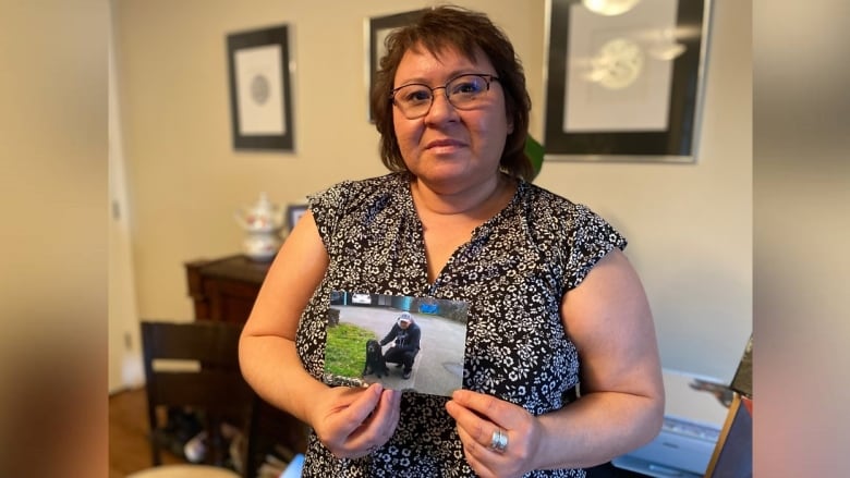 Women wearing glasses standing while holding a photo of her son in front of her.