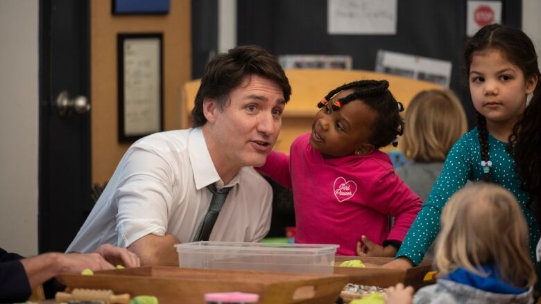 A man in a white shirt and tie neals next to a child wearing a pink shirt in front of what appears to be an arts and crafts table.