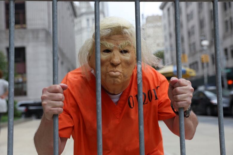 Man in Trump mask and orange prison jumpsuit, behind makeshift bars