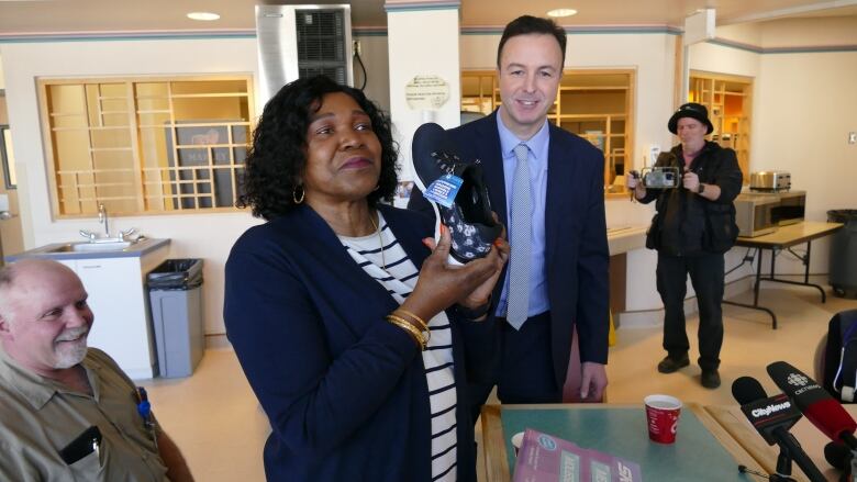 A woman is pictured holding up a black shoe to the camera as a man stands next to her, smiling.