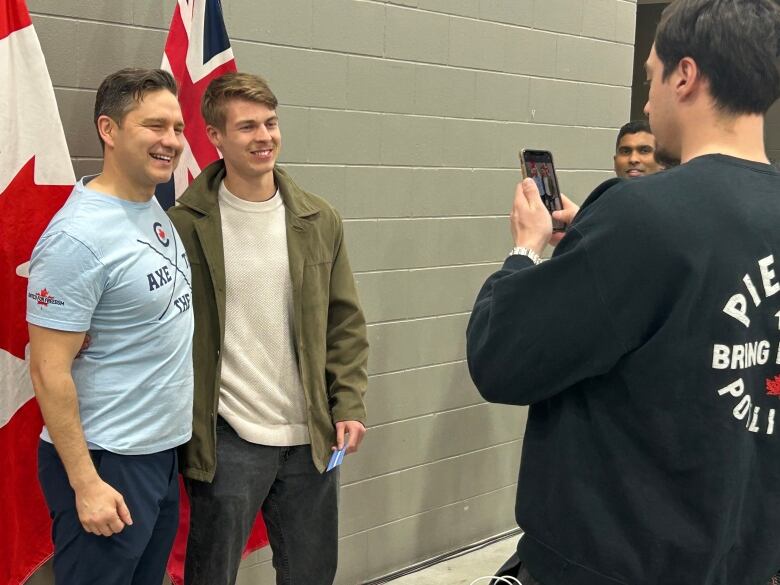 A man in a light blue t-shirt stands beside a man in a white sweater and green top as they pose together for a photo.