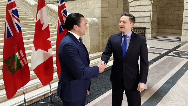 Two men in suits shake hands at the base of a staircase in front of Manitoba and Canadian flags