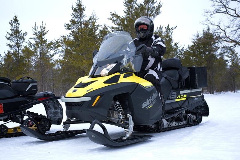 A man in a black snowsuit sits atop a black and yellow snowmobile in the winter in the woods.