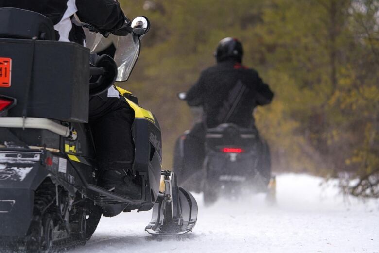 Two men on snowmobiles can be seen from behind as they travel down a snowy trail.