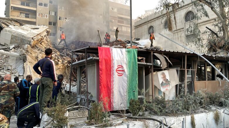 An Iranian flag hangs as smoke rises from a collapsed building.