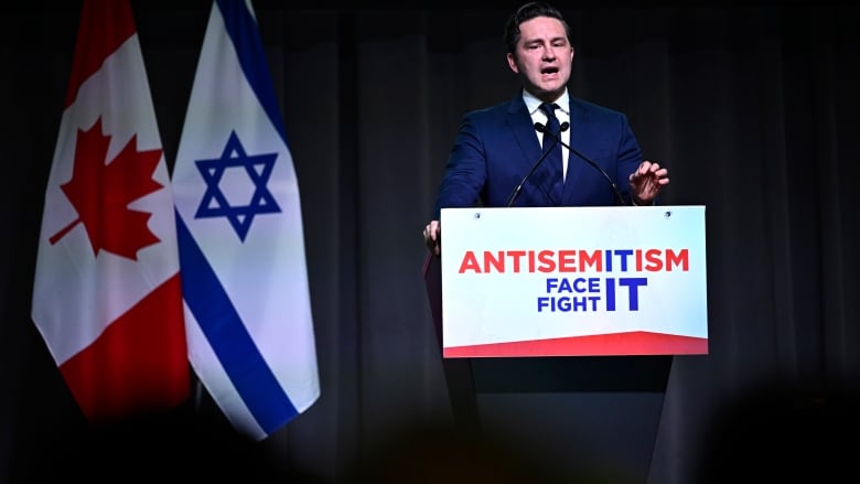 A man in a blue suit speaks at a podium, with the flags of Canada and Israel nearby.