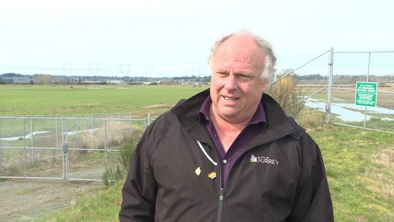 A white man wearing a City of Surrey jacket talks in front of a fence.