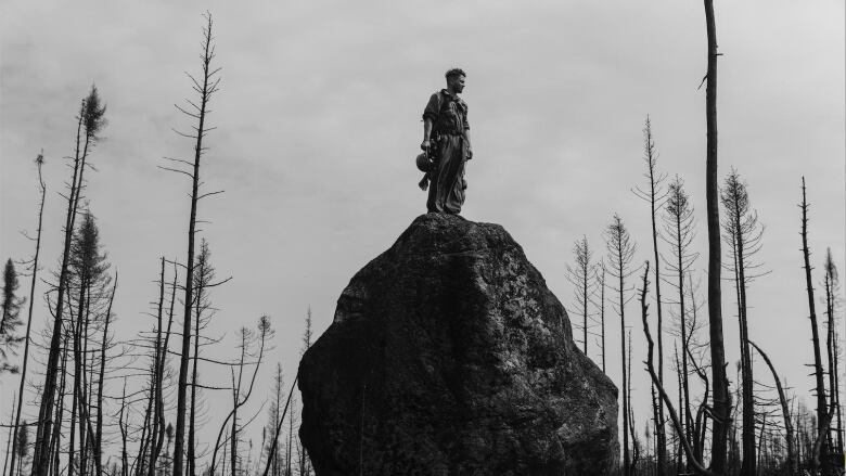 A wildfire fighter stands on a tall boulder amid charred trees.