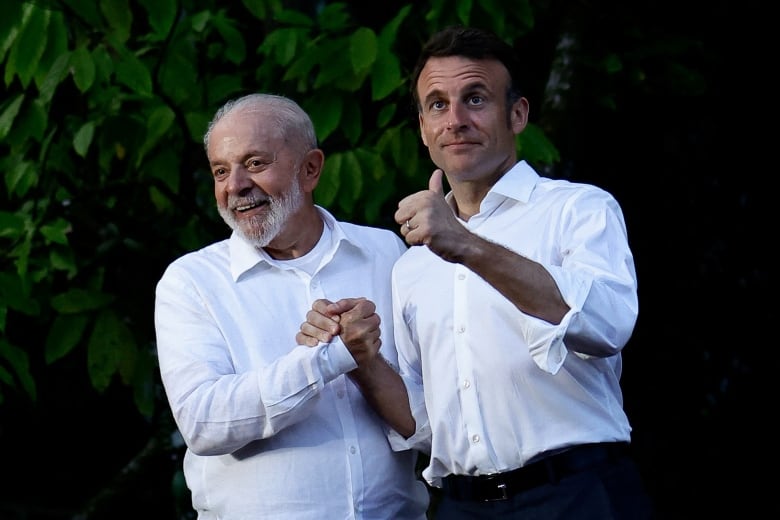 Two men wearing white button-down shirts grasp hands near some trees. One also gives a thumbs up.