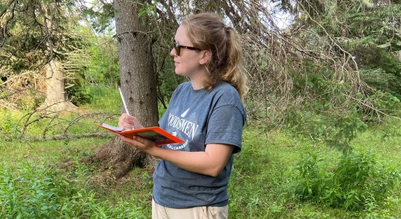 A young woman with a ponytail and sunglasses stands in nature, holding a pen and notebook. 