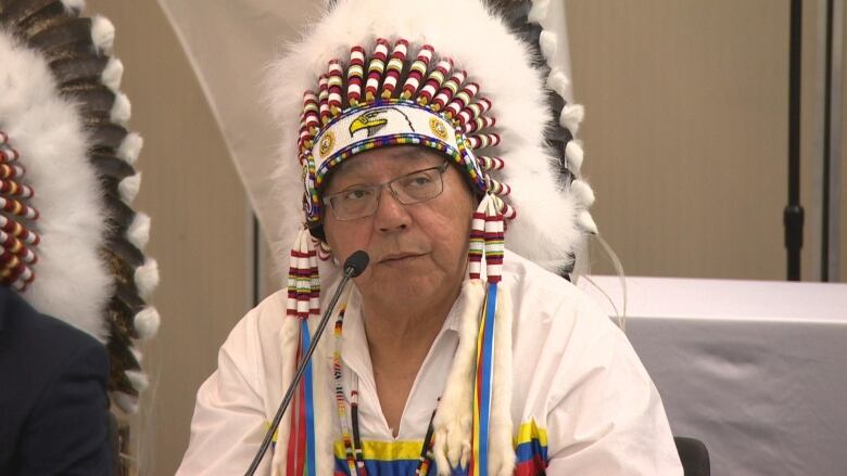 A man sits at a table with a microphone wearing a traditional headdress.