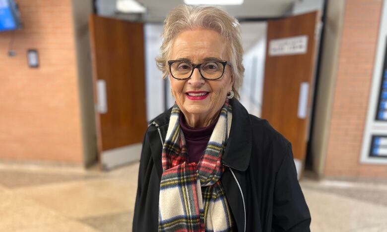 Older woman with short white hair and glasses stands in a school hallway, wearing a black coat and red, black and white plaid scarf.