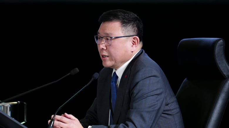 A man wearing a suit sits at a table speaking into a conference microphone