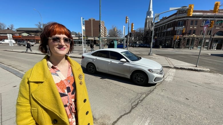 A woman wearing sunglasses and a yellow coat is standing on a street corner. A white car is making a right turn behind her at a wide intersection.