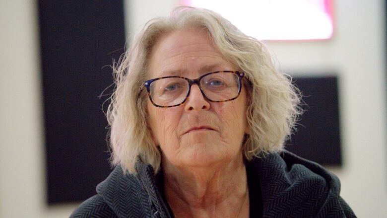 A woman with shoulder-length grey hair and mottled brown frame glasses looks into the camera.