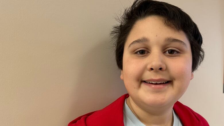 A young boy with brown hair, a white t-short and a red button up shirt layered over top smiles for the camera. He stands in front of an off-white wall. 