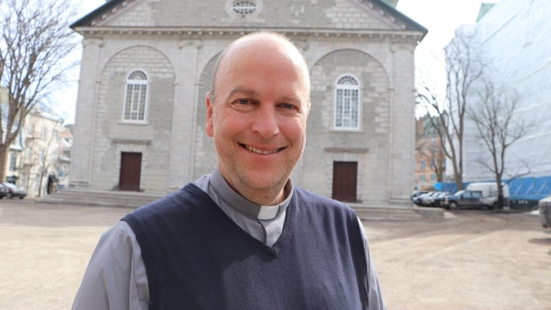 A man smiles at the camera standing in front of a church wearing a clerical collar.