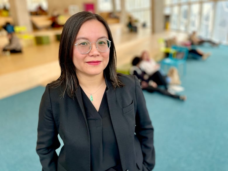 A woman wearing glasses and a black jacket stands with her hands in her pocket.