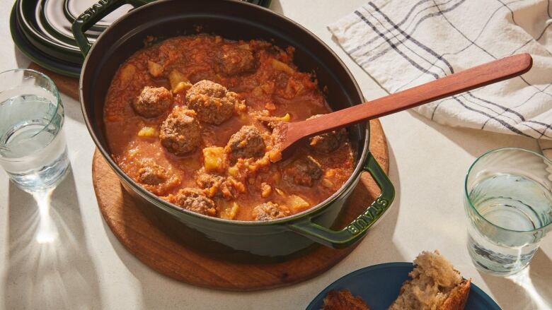 Overhead shot of dark green Dutch oven with a batch of Sweet-and-Sour Meatballs in it. Chunks of pineapple are visible. A wooden spoon sits in the dish. It's sitting on a wooden cutting board on a beige countertop with water glasses and a kitchen towel. 