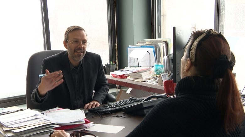 A man wearing glasses sits opposite a woman at a desk with large windows behind him.