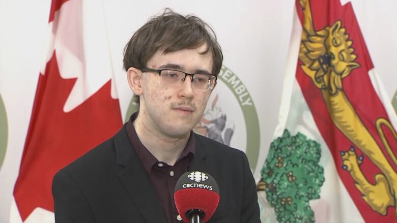 A young man in black and glasses speaks in front of a PEI flag.