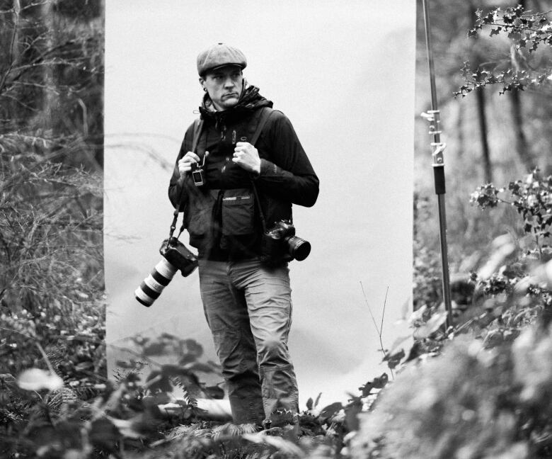 A man stands in a forest holding multiple cameras.