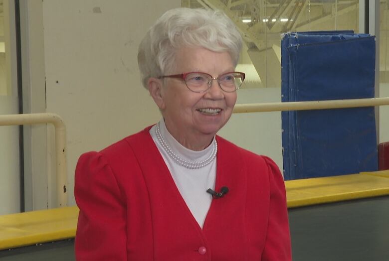 A woman with white hair sits, smiling, in a gymnastics club.