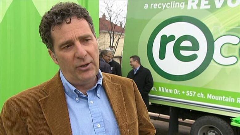 A man in a brown corderoy jacket and blue shirt stands in front of a truck with a bright grteen logo on the side.