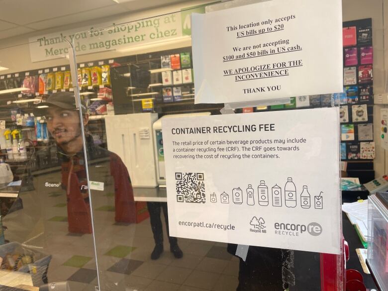 A clerk stands in the window of a Needs convenience store. Papers taped to the window advertize new recycling fees.