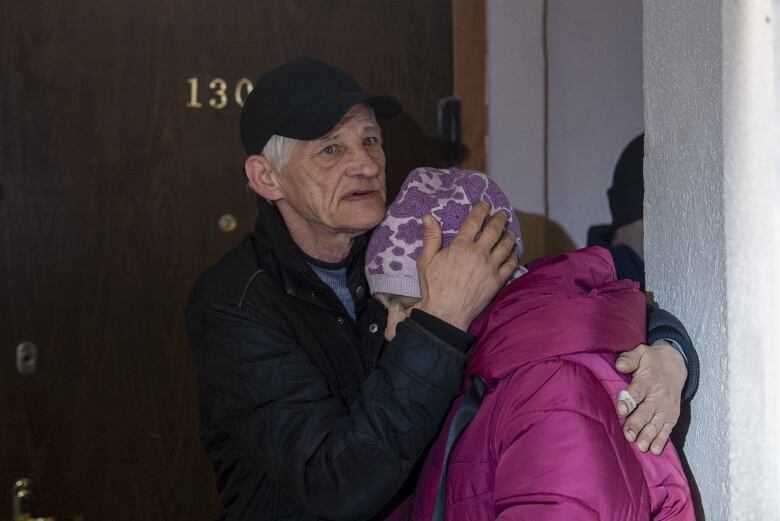 A man wearing a black cap hugs a woman wearing a pink coat inside a building.