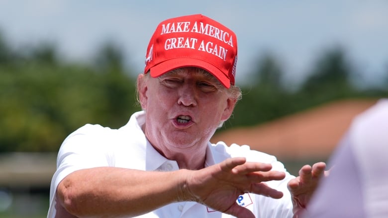 A cleanshaven older man in a baseball cap and white collared shirt is shown gesturing with his hands in an outdoor setting.