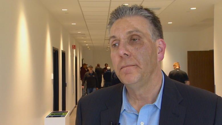 A man stands in front of the camera, wearing a suit and a blue shirt, with a long hallway behind him.