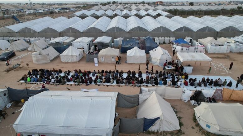 People sit on a long white blanket surrounded by tents