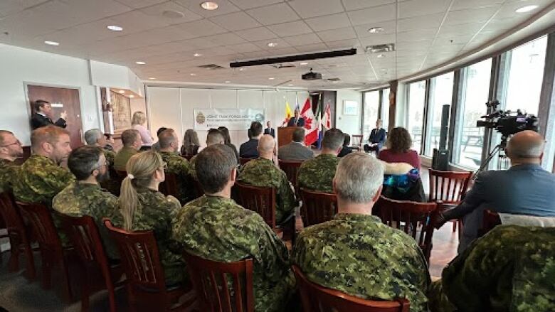 Military personnel dressed in green camo are seen from behind watching a man speak at a podium.