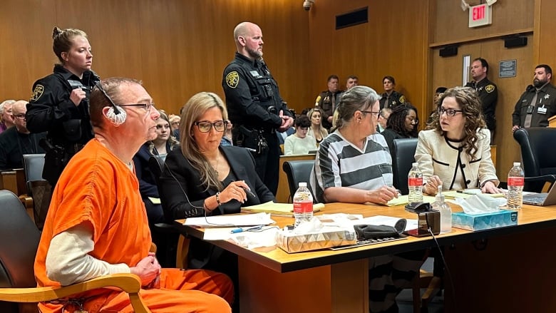 A man and a woman in prison uniform are shown seated at a courtroom table, with two women in professional attire shown beside them.