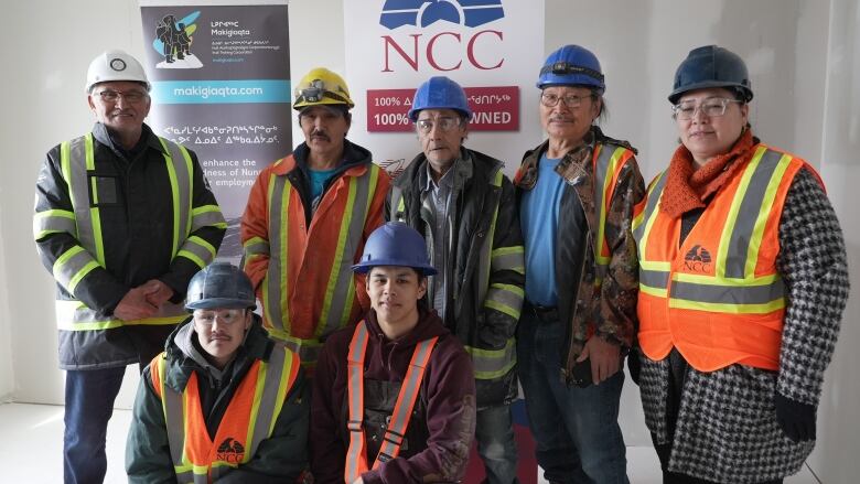 Seven people wearing orange and yellow vests and construction hats pose for a photo. 