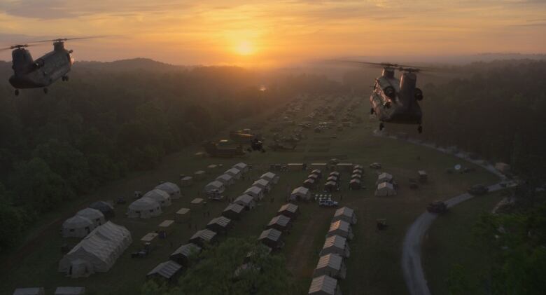 Two large army helicopters arrive a gathering point, a large field filled with military equipment and tents. 