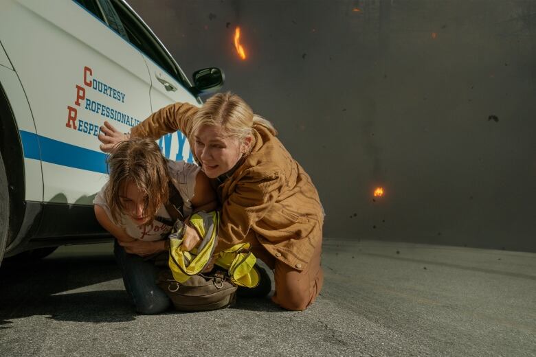 two women hide behind a police car as fires burn