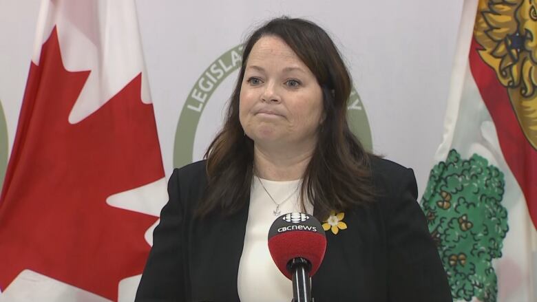 A woman in a black blazer and white shirt stands in front of a microphone taking questions from reporters.