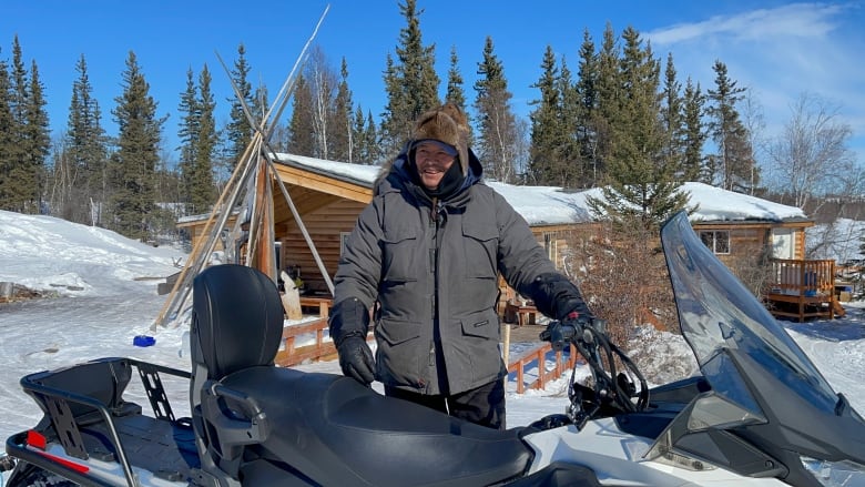 A man in a parka stands near a snowmobile in front of a log cabin, in winter.