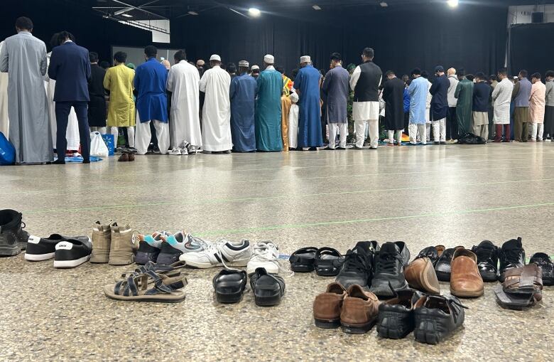 Shoes are seen lined up in the foreground with a line of people seen from behind, standing shoulder-to-shoulder, in the background.