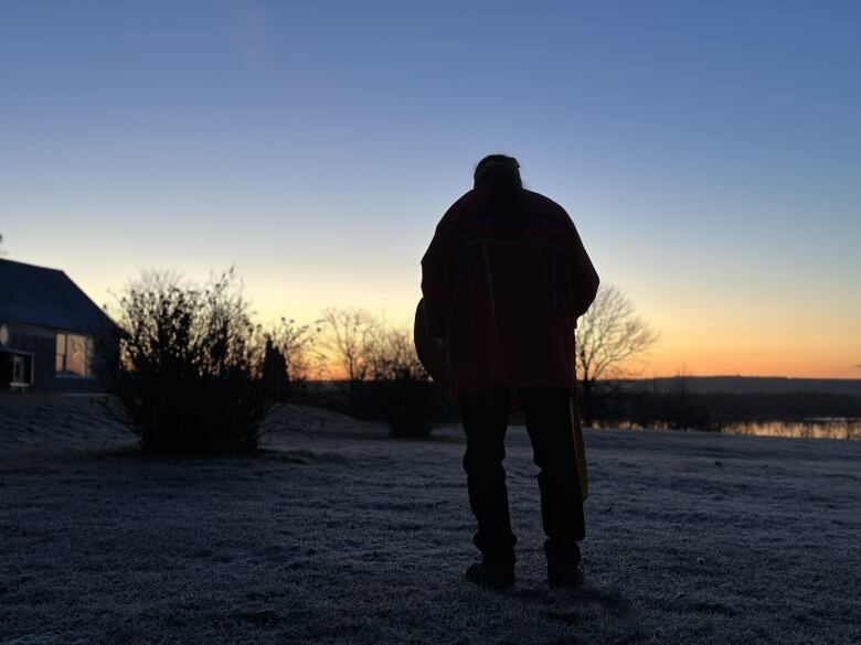 A man stands on grass facing the horizon, where the sun is setting.