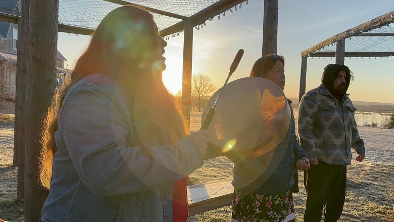 A woman with long dark hair glowing in the sunlight holds up a hand drum.