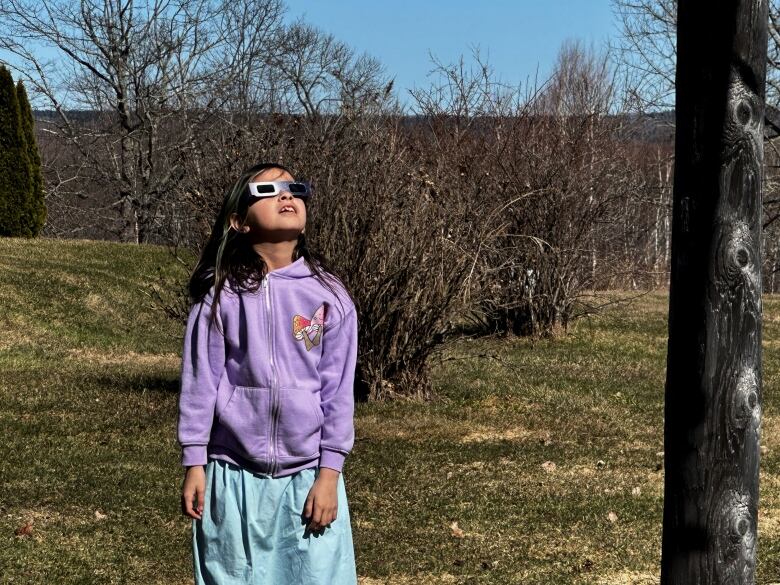 A young girl with long dark hair and wearing a purple sweater and light blue skirt stands outside staring up at the sun. She's wearing dark shades.